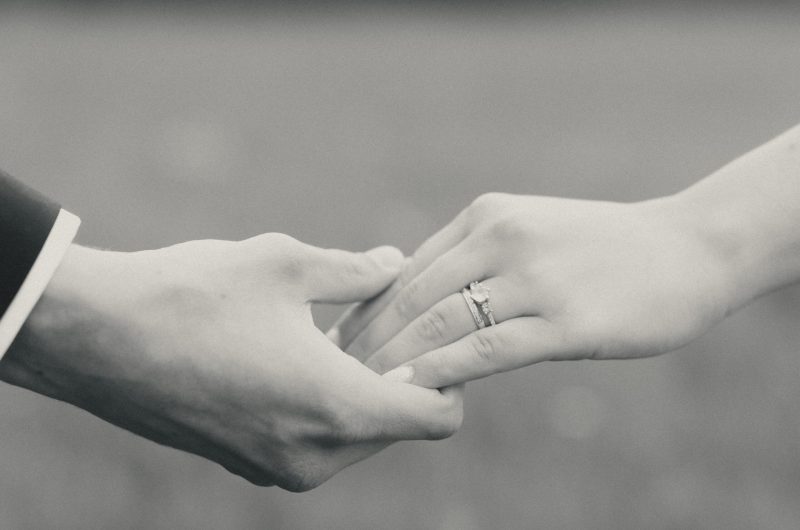A close-up photo on a bride and her groom holding hands with their wedding rings visible.
